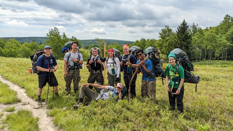 A group of hikers poses in a grassy field with backpacks. Some are standing, while one is lying down. The hikers are surrounded by trees and a cloudy sky. They appear to be taking a break during their hike.